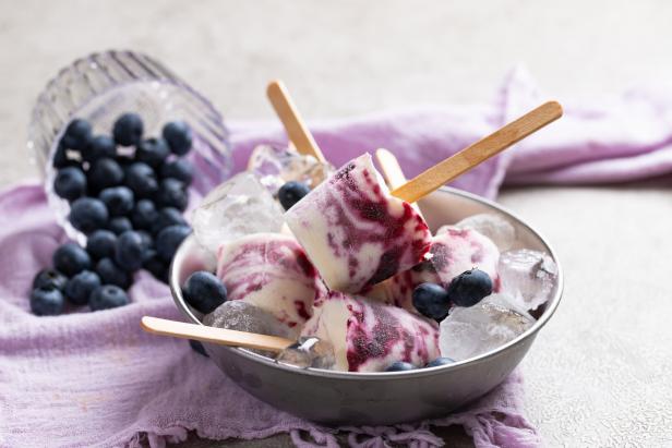 In einer Schale liegen Heidelbeer-Eis am Stiel mit Eiswürfeln und frischen Heidelbeeren auf einem violetten Tuch.