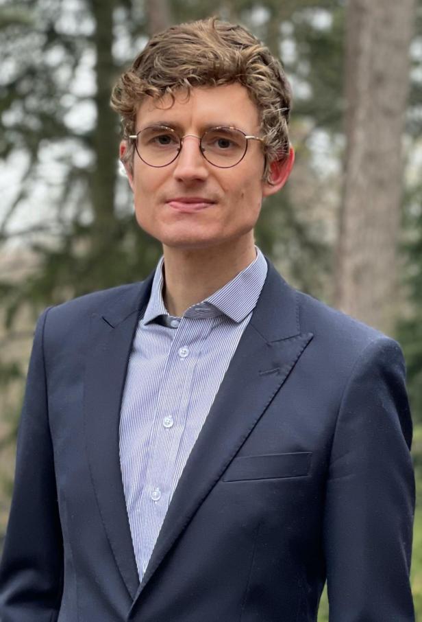 Ein Mann mit lockigen Haaren, Brille und Anzug steht vor unscharfem, bewaldetem Hintergrund.