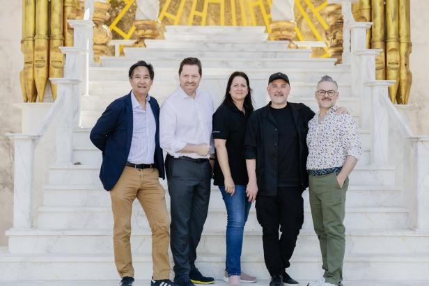 Fünf Personen stehen lächelnd nebeneinander auf einer weißen Marmortreppe mit goldenen Verzierungen im Hintergrund.
