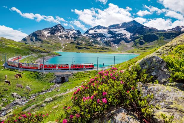 Ein roter Zug fährt vorbei an Bergen und einem türkisen See 