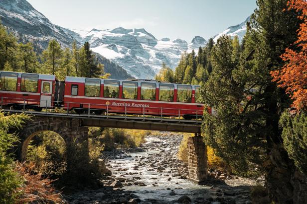 Der Bernina Express fährt über eine Brücke vor schneebedeckten Alpen und herbstlichen Bäumen.