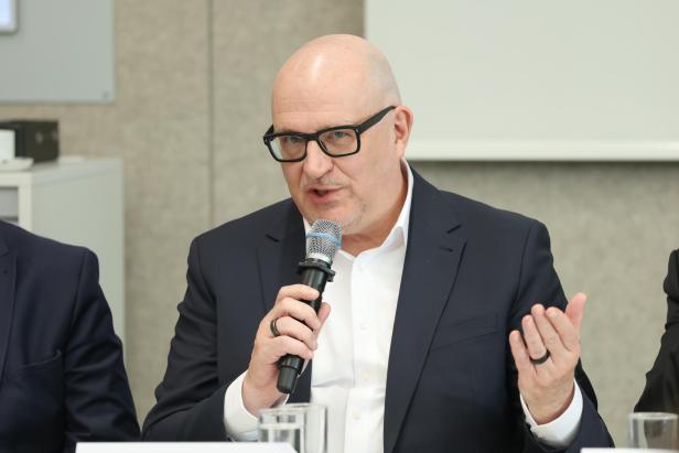 Ein Mann mit Brille und Anzug spricht in ein Mikrofon und gestikuliert mit der freien Hand.