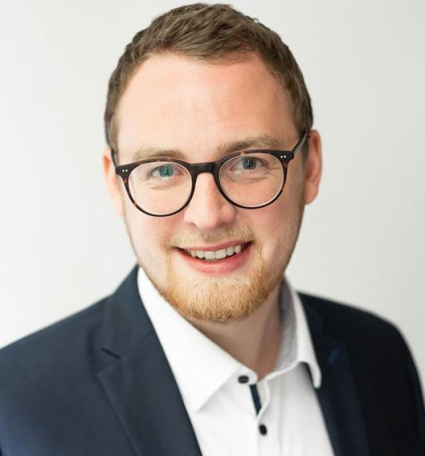 Mann mit Brille und Bart im Anzug vor neutralem Hintergrund.