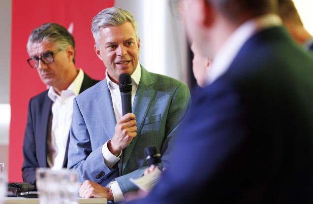 Ein Mann im blauen Anzug spricht mit einem Mikrofon, weitere Personen stehen und sitzen um ihn herum.