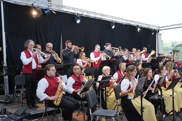 Eine Blaskapelle mit Musikern in Tracht spielt auf einer Bühne vor schwarzem Hintergrund.