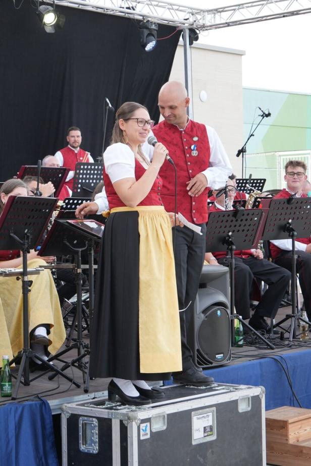 Eine Frau und ein Mann in Tracht stehen auf einer Bühne vor einem Blasorchester, die Frau spricht ins Mikrofon.
