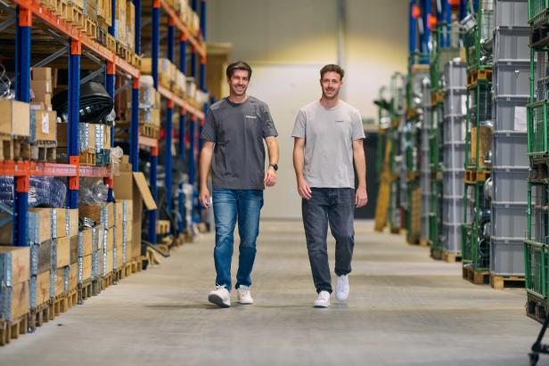 Zwei Männer in T-Shirts mit dem Logo von irriport24 gehen durch einen Lagergang zwischen hohen Regalen mit Waren.