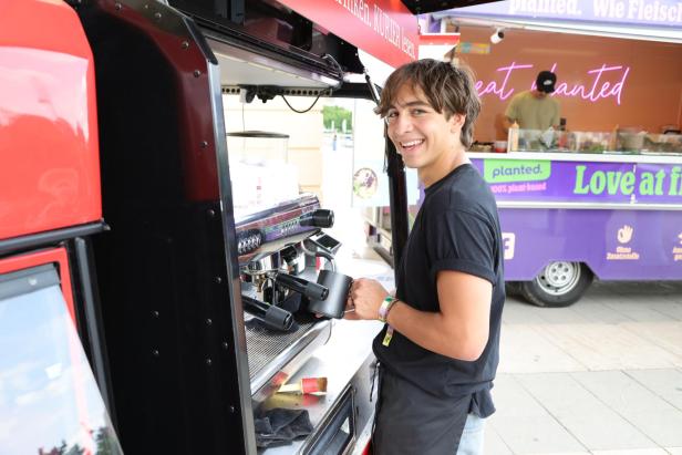 Ein junger Mann bereitet an einer Kaffeemaschine in einem mobilen Café einen Kaffee zu und lächelt dabei freundlich.