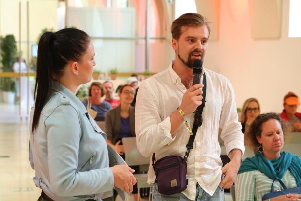 Ein Mann spricht mit einem Mikrofon, während eine Frau ihm zuhört und mehrere Personen im Hintergrund sitzen.