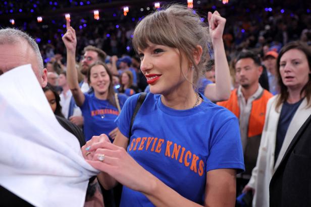 Taylor Swift trägt ein blaues T-Shirt mit der Aufschrift „Stevie Knicks“ und lächelt in einer Menschenmenge.