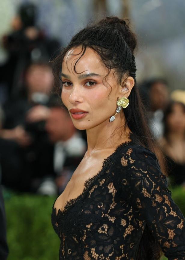 Zoë Kravitz trägt ein schwarzes Spitzenoberteil und auffällige goldene Ohrringe vor einem unscharfen Hintergrund.