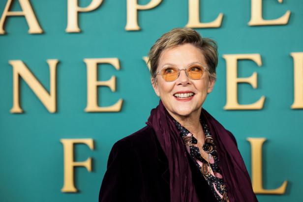 Annette Bening lächelt vor einem türkisfarbenen Hintergrund mit goldener Schrift und trägt eine Brille mit getönten Gläsern sowie einen dunklen Schal.