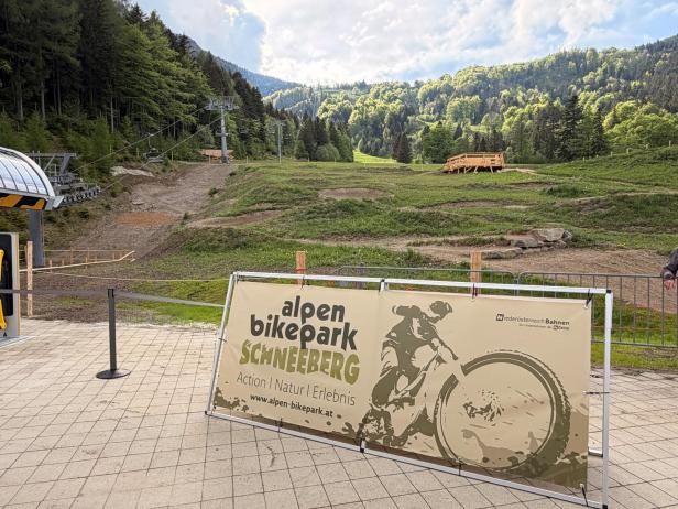 Eingangsbereich des Alpen Bikepark Schneeberg mit Werbebanner, Blick auf die Mountainbike-Strecken und die umgebende grüne Berglandschaft.