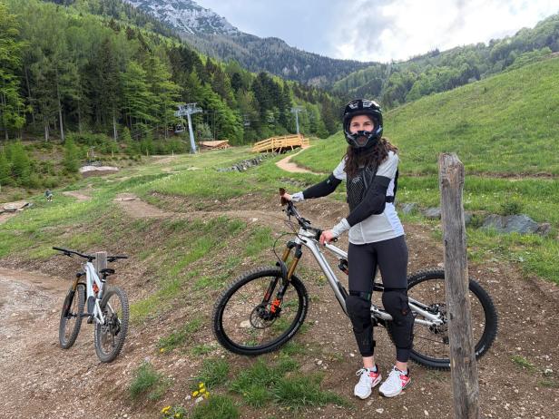 Frau mit Helm, Knieschützern und Sportkleidung steht neben Mountainbike auf einem grasbewachsenen Weg in bergiger Landschaft mit Wald und Sessellift im Hintergrund.