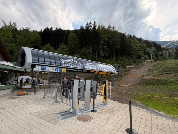 Bergstation einer Sesselliftanlage mit Warteschlangenabsperrungen vor bewaldetem Hang und bewölktem Himmel.