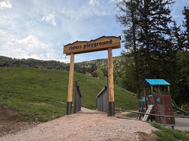 Holzportal mit der Aufschrift 'riders playground' am Eingang zu einem Spielplatz mit Klettergerüst vor bewaldetem Hügel.