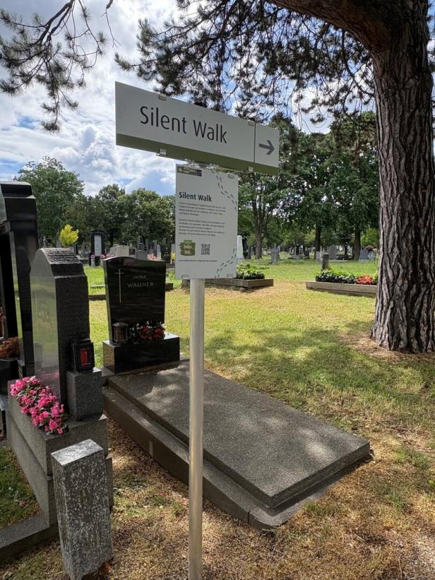 Wegweiser mit der Aufschrift „Silent Walk“ auf einem Friedhof, umgeben von Grabsteinen und Bäumen.