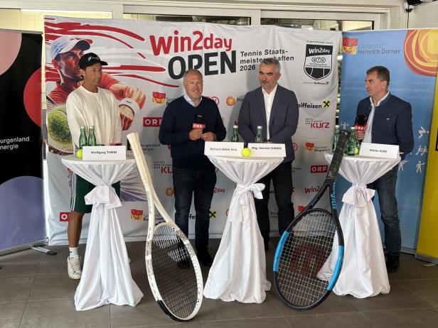 Vier Männer stehen an Stehtischen mit Namensschildern und riesigen Tennisschlägern bei einer Tennisveranstaltung.