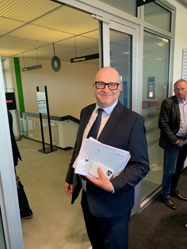 Johannes Larcher im Anzug steht lächelnd mit Unterlagen und Smartphone in der Hand vor einem modernen Büroflur.