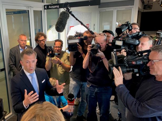 Ein Mann im Anzug spricht vor mehreren Kameraleuten und Journalisten, die ihn mit Mikrofonen und Kameras umgeben.