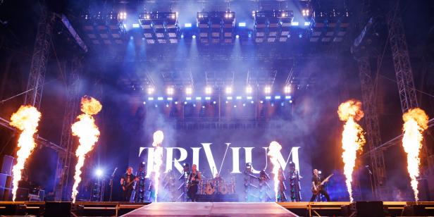 Mehrere Musiker treten auf einer großen Bühne mit Pyrotechnik und dem Schriftzug „TRIVIUM“ im Hintergrund auf.