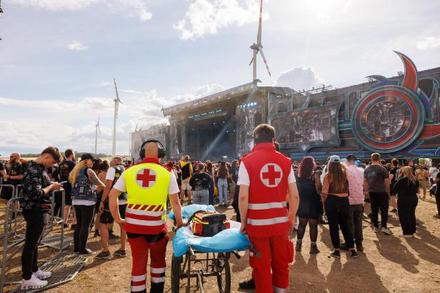 Zwei Sanitäter mit Trage stehen bei einem Open-Air-Konzert vor einer großen Bühne und beobachten die Menschenmenge.