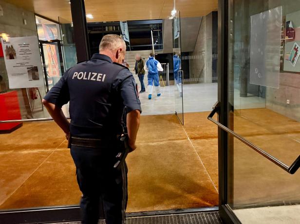 Ein Polizist steht vor einem Eingang, im Hintergrund gehen Personen in Schutzanzügen durch einen modernen Flur.