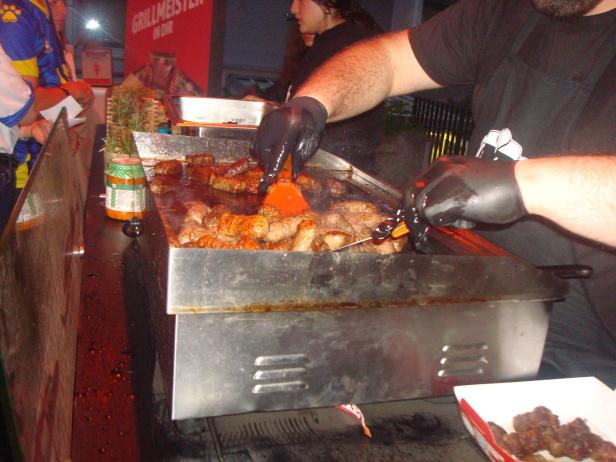 Grillmeister bereitet Ćevape auf einer heißen Grillplatte vor, daneben warten Gäste in Trikots.