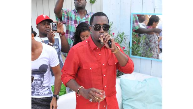 P. Diddy raucht eine Zigarre und hält ein Glas Wein in der Hand, umgeben von Menschen.