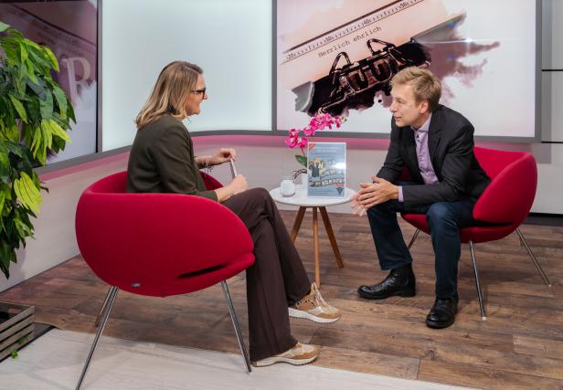 Zwei Personen sitzen sich in roten Sesseln gegenüber und unterhalten sich, auf dem Tisch steht eine Orchidee und ein Buch.
