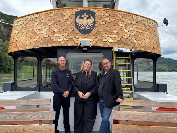 Drei Personen stehen auf einem Schiff vor einer Kabine mit goldfarbenen Schuppen und einem Wappen mit Doppeladler.