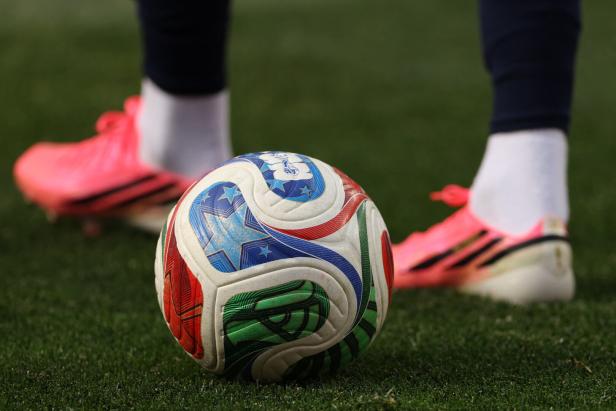 Ein bunter Fußball liegt auf Rasen zwischen zwei Füßen in pinken Fußballschuhen.