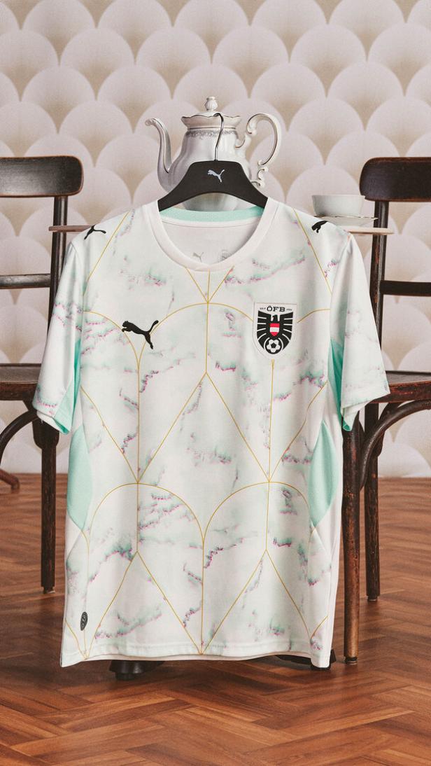 Ein weißes Fußballtrikot mit mintgrünen Akzenten, goldenen Linien und dem österreichischen Verbandswappen hängt auf einem Stuhl.