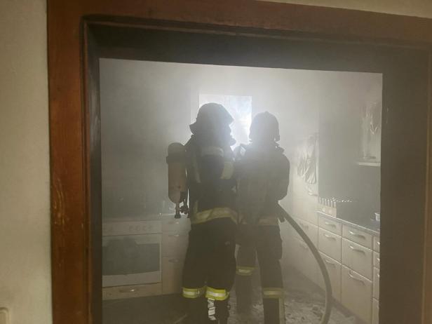 Zwei Feuerwehrleute mit Atemschutzgeräten bekämpfen Rauch und Feuer in einer verrauchten Küche.