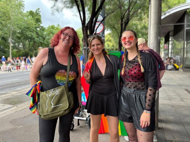 Drei Frauen mit Regenbogen-Accessoires und fröhlichen Gesichtern stehen auf einer Straße, im Hintergrund sind Bäume und weitere Menschen.