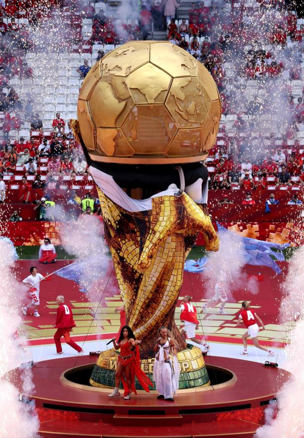 Eine große goldene Trophäe steht im Stadion, umgeben von Tänzern, Rauch und Funken bei einer festlichen Show.
