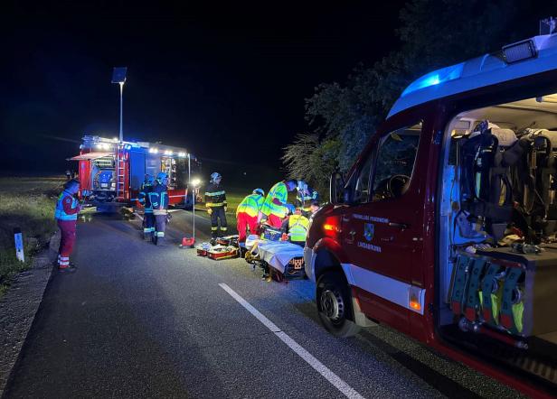 Mehrere Einsatzkräfte versorgen nachts eine verletzte Person auf einer Landstraße, umgeben von Rettungs- und Feuerwehrfahrzeugen.
