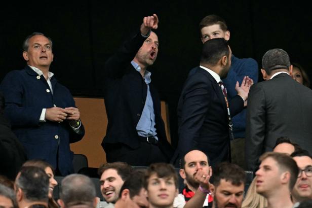 Prinz William gestikuliert aufgeregt und zeigt mit dem Finger auf das Spielfeld, während er mit anderen Personen auf der Tribüne sitzt.