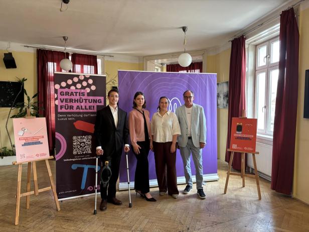 Vier Personen stehen vor einem violetten Banner für die Initiative „Gratis Verhütung für alle“ mit flankierenden Infotafeln auf Staffeleien.