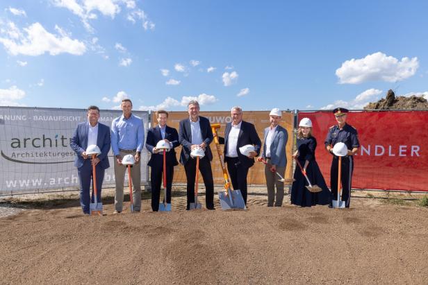 Mehrere Personen stehen mit Schaufeln und Helmen auf einer Baustelle bei einer offiziellen Spatenstich-Zeremonie.