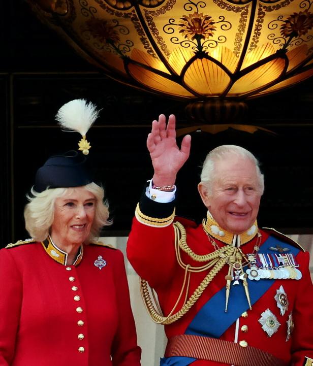 König Charles III. und Königin Camilla winken in festlichen Uniformen von einem Balkon. Charles' Armbändchen ist zu sehen.