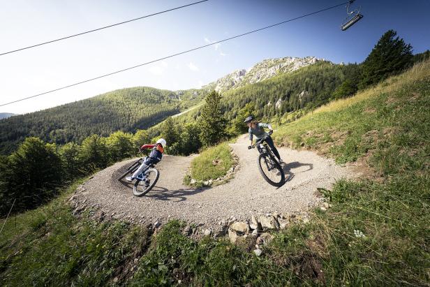Zwei Mountainbiker fahren auf einer kurvigen Trail-Abfahrt im Alpen Bikepark Schneeberg vor alpiner Bergkulisse.