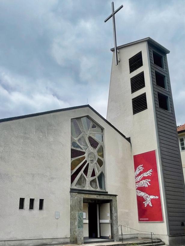 Kirche mit Kreuzturm, rundem Glasfenster und rotem Wandbild unter wolkigem Himmel.