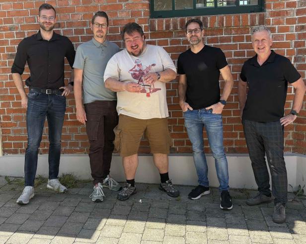 Fünf Männer stehen lächelnd vor einer roten Backsteinwand.