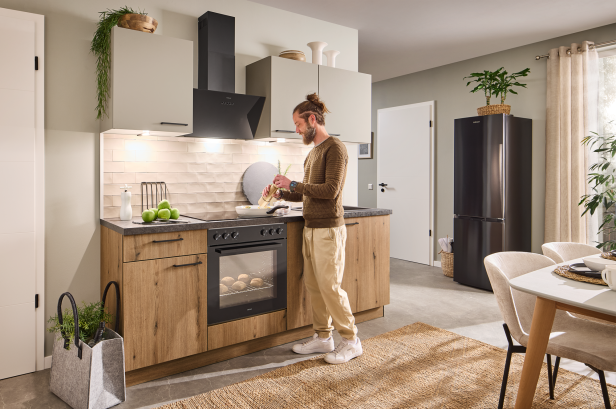 Kleine Küchenzeile mit Holzfronten, schwarzem Backofen und Dunstabzugshaube, daneben steht eine Person, rechts ein schwarzer Kühlschrank und ein Esstisch mit Stühlen
