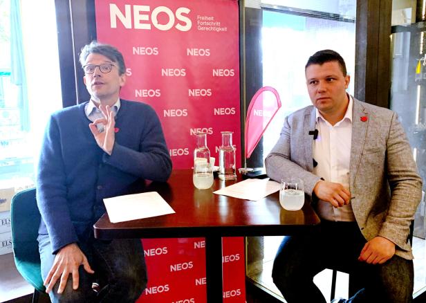 Zwei Männer sitzen vor einem roten NEOS-Banner an einem Tisch und führen ein Gespräch.