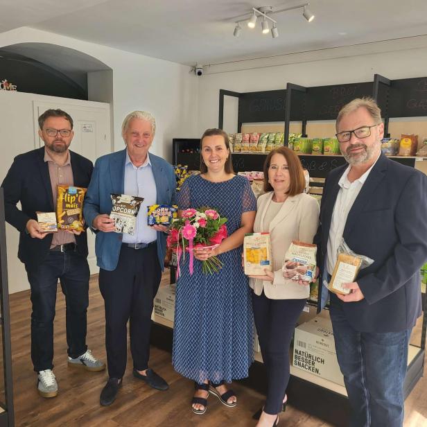 Fünf Personen stehen in einem Laden und halten verschiedene Snacks, die Frau in der Mitte ein Blumenstrauß.