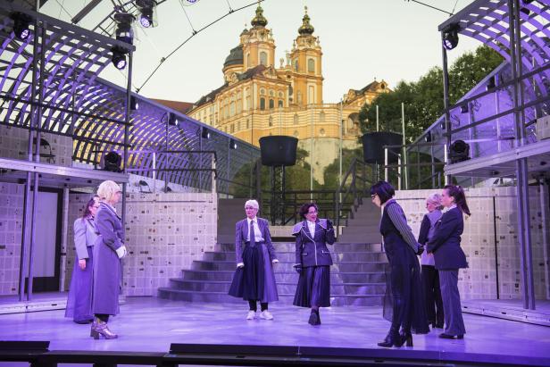Mehrere Personen stehen auf einer Bühne mit violetter Beleuchtung vor einer barocken Kirche im Hintergrund.