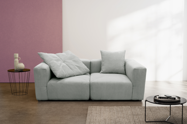 Hellgraues Sofa mit zwei Kissen vor einer weißen Wand und einem großen rosa Bild, daneben zwei runde Beistelltische mit Dekoration