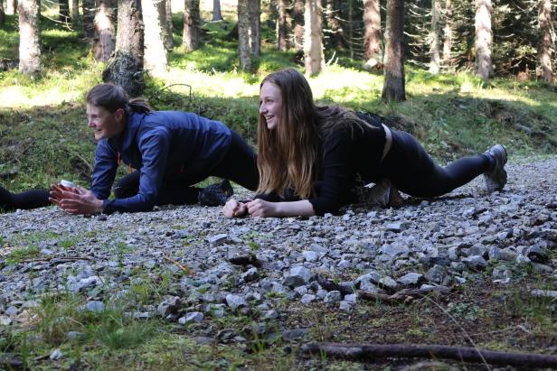 Zwei Frauen machen im Wald eine Dehnübung auf einem Kiesweg.